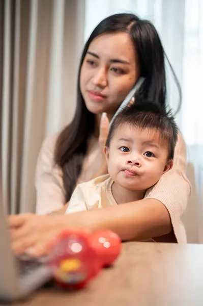 A busy Asian businesswoman mom is multitasking, talking on the phone with her client, focusing on her laptop, and taking care of her little son at home. work from home and mom\'s life concepts