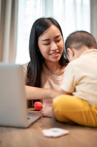 A happy Asian mom is talking and playing with her little son while working on her laptop computer, working remotely at home.