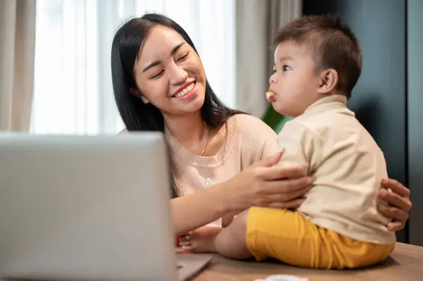 A happy Asian mom is playing and feeding her little son with snacks while working in her home office. work from home and mom\'s life concepts
