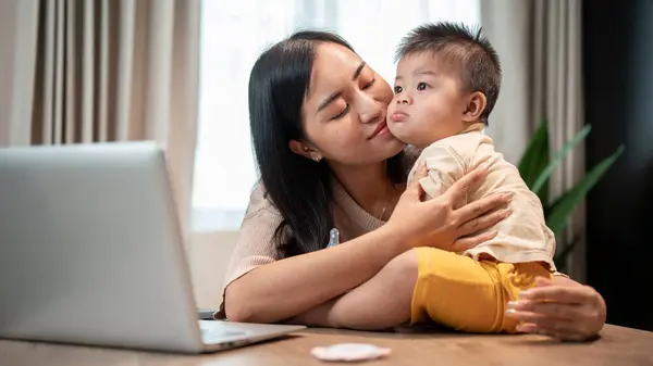 A happy Asian mom is playing and kissing her cute little son while working in her home office. family bonding, mom\'s life, happy motherhood