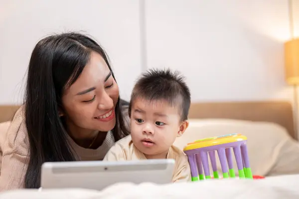 A happy Asian mom and her little son are watching a kid\'s cartoon on a digital tablet together in bed in the bedroom. motherhood, mom\'s life, family bonding