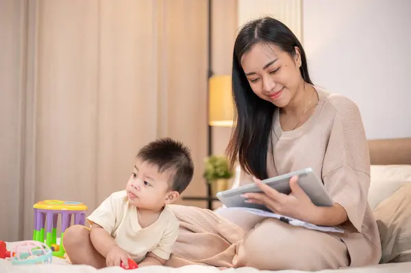 A beautiful Asian mom is working on her digital tablet while her bay boy is playing toys in bed. mom\'s life concept