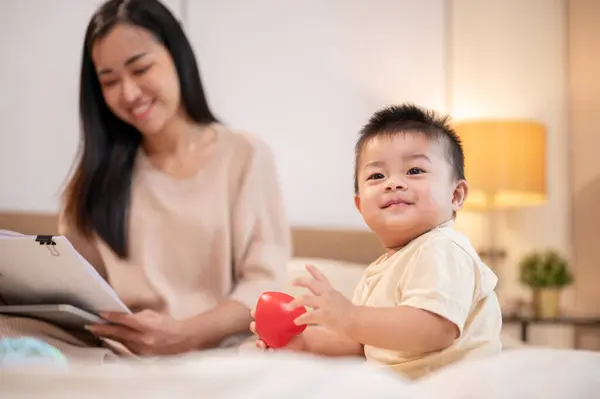 A happy, cute Asian baby boy is playing with toys while his mom is working in bed, spending time together in the bedroom. toddler, baby, kid, childhood