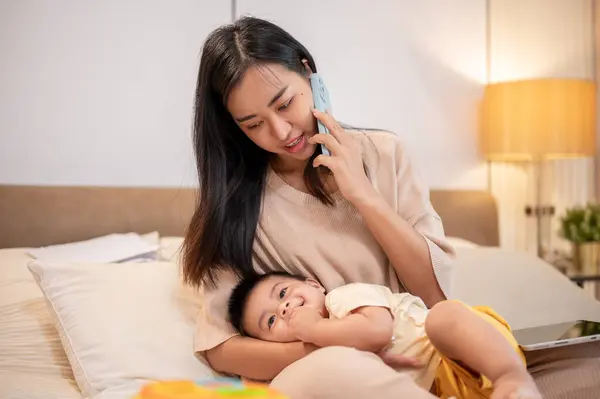 A caring Asian mom is putting her little son to sleep in her arms while she is talking on the phone with someone. napping and bedtime, motherhood and childhood