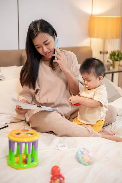 Young busy Asian mother is reading documents and talking on the phone with someone while taking care of her baby boy in bed in the bedroom. businesswoman mom, motherhood, busy mom\'s life, single mom