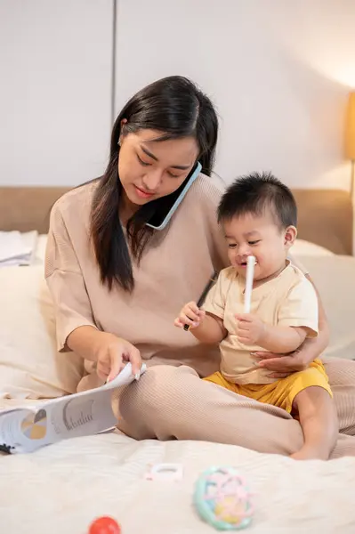 A busy young Asian mom is taking care of and holding her little baby boy while talking on the phone and working on documents in bed in the bedroom. work from home and mom\'s life concepts