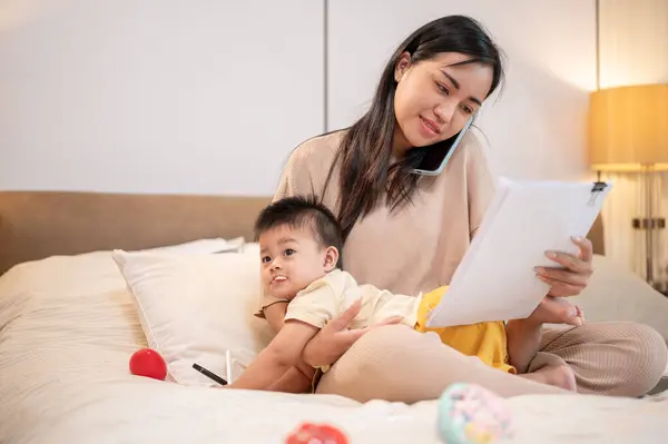 Young busy Asian mother is reading documents and talking on the phone with someone while taking care of her baby boy in bed in the bedroom. businesswoman mom, motherhood, busy mom\'s life, single mom