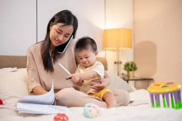 A busy young Asian mom is taking care of and holding her little baby boy while talking on the phone and working on documents in bed in the bedroom. work from home and mom's life concepts
