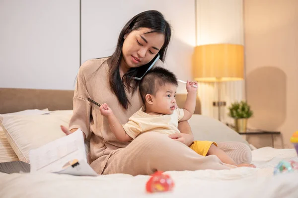 A busy young Asian mom is taking care of and holding her little baby boy while talking on the phone and working on documents in bed in the bedroom. work from home and mom\'s life concepts