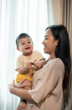 Olumlu ve ilgili bir Asyalı anne, sevimli bebeğini oturma odasının penceresinin yanında oynatıyor ve kucağına alıyor. annelik ve çocukluk kavramları