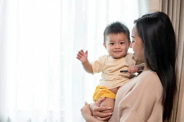 A positive and caring Asian mom is playing and holding her cute baby boy in her arms near by the window in the living room. motherhood and childhood concepts