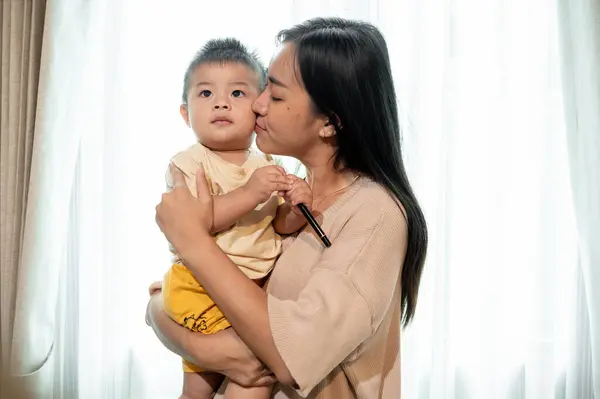 A loving and caring Asian mom is kissing and holding her cute little son in the living room. motherhood and childhood concepts