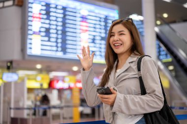 Neşeli bir Asyalı bayan yolcu havaalanında dururken elini sallıyor ve arkadaşlarını arıyor. seyahat, tatil, tatil