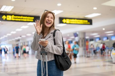 Neşeli ve gülümseyen Asyalı bir bayan yolcu havaalanında dururken elini sallıyor ve arkadaşlarını arıyor. seyahat, tatil, tatil