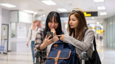 İki Asyalı bayan arkadaş havaalanında bir bagaj vagonuyla yürürken uçuş bilgilerini ve uçuş saatlerini kontrol ediyorlar..
