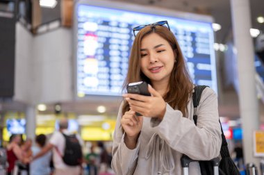 Güzel, mutlu bir Asyalı kadın havaalanında dikilirken akıllı telefonunu kullanıyor, sohbet ediyor ve mesajları kontrol ediyor..