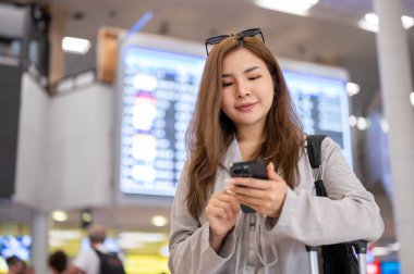 Güzel, mutlu bir Asyalı kadın havaalanında dikilirken akıllı telefonunu kullanıyor, sohbet ediyor ve mesajları kontrol ediyor..