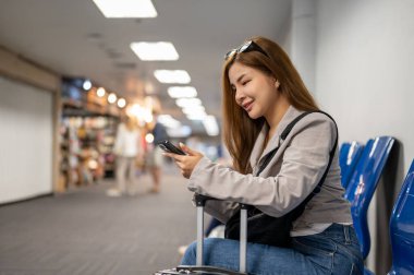 Güzel bir Asyalı bayan yolcu, havaalanı terminalinde beklerken arkadaşlarıyla sohbet etmek için akıllı telefonunu kullanıyor..