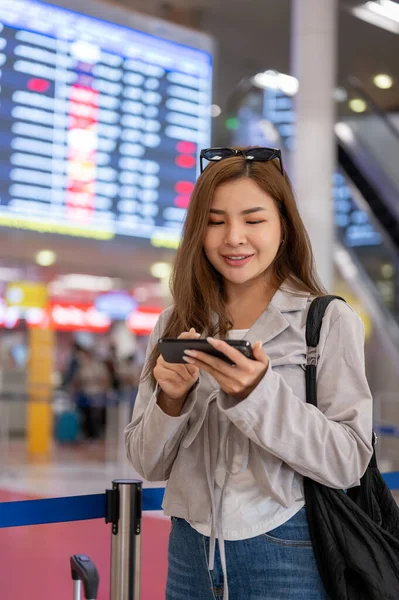 An attractive Asian female passenger is using her smartphone to check her flight boarding time while standing in front of a boarding time monitor in the airport. people and transportation concepts
