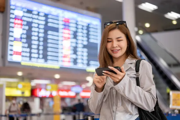 An attractive Asian female passenger is using her smartphone to check her flight boarding time while standing in front of a boarding time monitor in the airport. people and transportation concepts