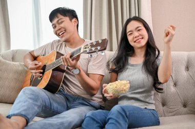 Neşeli bir Asyalı erkek arkadaş akustik gitar çalıp şarkı söylerken, kız arkadaşı da kanepede dans ediyor ve evde eğleniyor. ev hayatı ve çift kavramları