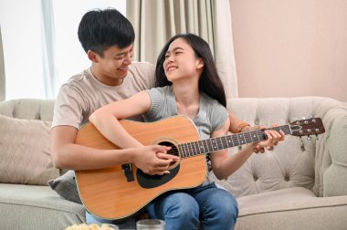 Mutlu genç bir Asyalı çift oturma odasında bir kanepede birlikte gitar çalıyor. Bir erkek arkadaş kız arkadaşına evde gitar çalmayı öğretiyor..