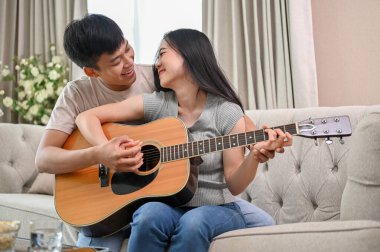 Mutlu genç bir Asyalı çift oturma odasında bir kanepede birlikte gitar çalıyor. Bir erkek arkadaş kız arkadaşına evde gitar çalmayı öğretiyor..