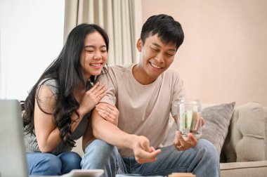 Genç ve mutlu bir Asyalı çift, oturma odasındaki bir kanepede tasarruf planlarından bahsederken kavanozda para biriktiriyor. insanlar ve finansal kavramlar