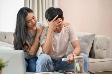İlgili genç bir Asyalı kadın, stresli eşini kanepede destekliyor ve teşvik ediyor. Genç Asyalı çift ev giderleri ve yüksek elektrik faturaları konusunda endişeli..
