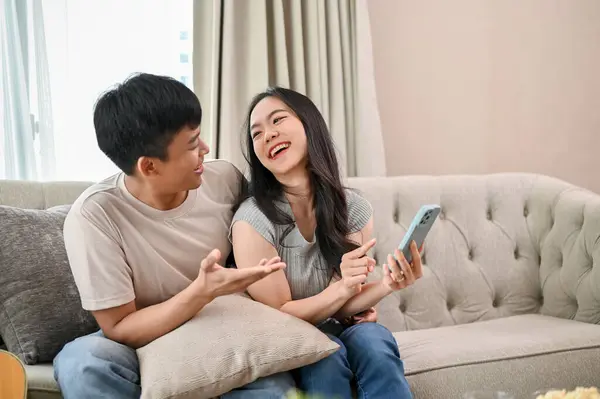 Happy young Asian couple, boyfriend and girlfriend, enjoy talking while using a smartphone together on a sofa in the living room.