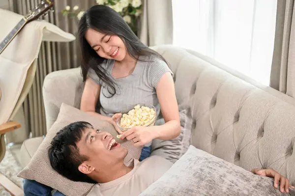 A happy young Asian man is lying on his girlfriend\'s lap on a sofa, sharing a cute and romantic moment at home together. couple, married couple, boyfriend and girlfriend