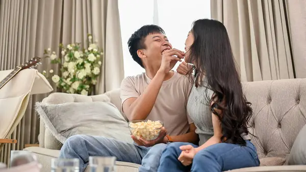 A cheerful and joyful young Asian couple is laughing at jokes and enjoying popcorn while watching television together at home. couple and domestic life concepts