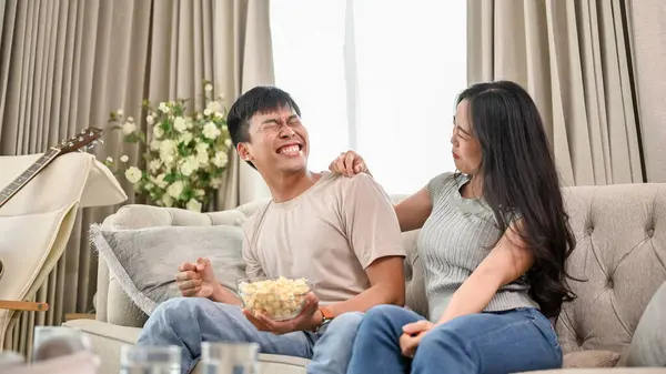 A cheerful and joyful young Asian couple is laughing at jokes and enjoying popcorn while watching television together at home. couple and domestic life concepts
