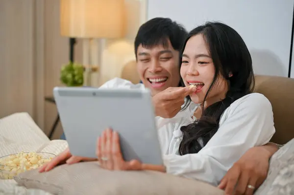 A cheerful and happy young Asian couple in pajamas are enjoying watching a movie on a digital tablet and eating popcorn in bed together. couple and domestic life concepts