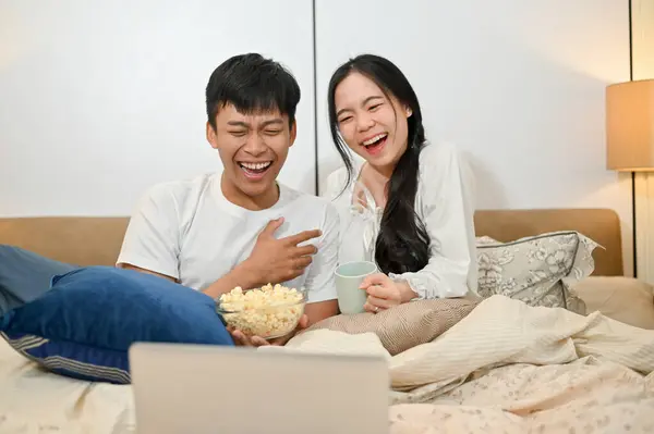 A cheerful and happy young Asian couple in pajamas is laughing, enjoying watching a movie on a laptop in bed, having a fun nighttime in the bedroom together. couple and bedtime concepts
