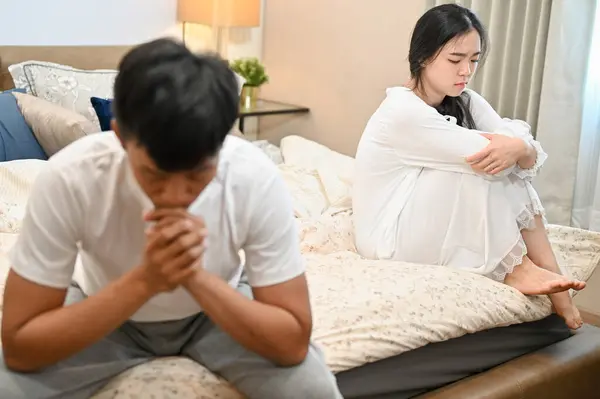 A sad, unhappy young Asian wife in pajamas is sitting separately with her husband on bed after their argument. relationship problems, misunderstanding, marriage conflict