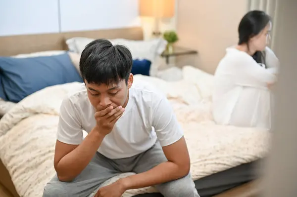 A sad, unhappy Asian husband in pajamas is sitting separately with his wife on bed after their argument. relationship problems, misunderstanding, marriage conflict