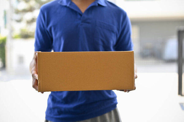 A cropped shot of a delivery man in a blue shirt holding a cardboard box in front of the door. package delivery, courier, postman, shipping