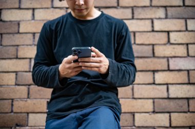 Havalı, şık giyinmiş havalı bir Asyalı 'nın kırpılmış görüntüsü, akıllı telefonunu sokakta tuğla bir duvarın yanında dururken kullanıyor. İnsanlar ve kablosuz teknoloji kavramları
