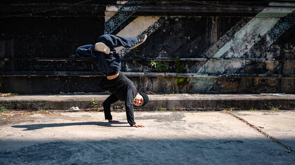 Холодный, веселый и профессиональный азиатский танцор хип-хопа практикует свой брейк-данс на заброшенной улице в солнечный день.