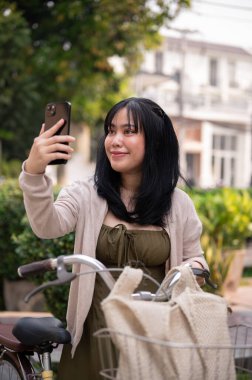 Şirin elbiseli, sevimli, mutlu bir Asyalı kadın akıllı telefonuyla şehrin fotoğrafını çekerken bisikletiyle geziyor. insanlar ve yaşam tarzı kavramları