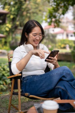 Sıradan giyinmiş mutlu bir Asyalı kadın arka bahçesinde dinlenirken akıllı telefonuyla arkadaşlarıyla sohbet etmekten zevk alıyor. İnsanlar ve kablosuz teknoloji kavramları
