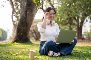 Parlak bir günde yeşil bir parkta dizüstü bilgisayarını kullanan mutlu bir Asyalı kadın. insanlar ve teknoloji kavramları