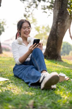 Güzel, mutlu Asyalı bir kadın akıllı telefonunu kullanıyor, çimlerin üzerinde oturuyor, güneşli bir günde yeşil bir parkta dinleniyor..