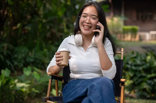 A positive Asian woman in casual clothes is sipping coffee and talking on the phone while relaxing in her backyard garden. people and lifestyle concepts