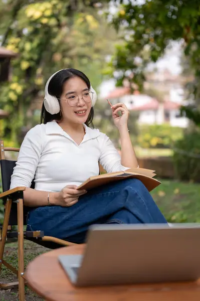 A happy and relaxed Asian woman is listening to music on her headphones, working or writing her diary while sitting in her green backyard garden.