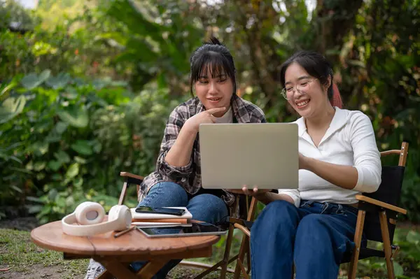 Two happy Asia female university students are discussing work and doing homework on a laptop together while sitting at a table in a backyard.