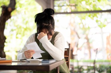 Asyalı genç bir bayan küçük işletme sahibi faturaları hesaplıyor, dükkanının giderleri ve gelirleri için endişeleniyor, finansal bir kriz yaşıyor ve aylık satışlarından hayal kırıklığına uğruyor..