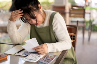 Asyalı genç bir bayan küçük işletme sahibi faturaları hesaplıyor, dükkanının giderleri ve gelirleri için endişeleniyor, finansal bir kriz yaşıyor ve aylık satışlarından hayal kırıklığına uğruyor..