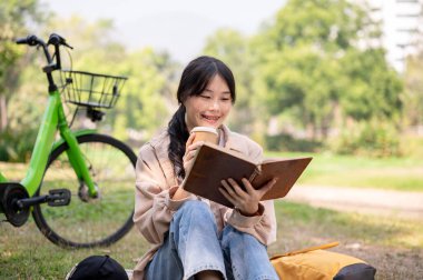 Olumlu, mutlu, genç Asyalı bir kadın yeşil bir parkta dinlenirken kitap okuyor ve kahvesini yudumluyor. insanlar ve yaşam tarzı kavramları